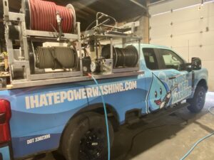 A blue truck with "Suds Power Washing" and a cartoon water droplet logo on the side is parked indoors. Equipment and hose reels are mounted in the truck bed, and "IHATEPOWERWASHING.COM" is printed on the side.