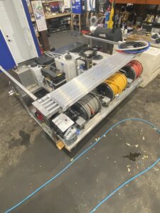 A cleaning equipment setup in a workshop, featuring multiple coiled hoses in red, yellow, and black, tanks, reels, and various supplies organized on a metal frame, with tools and spare parts in the background.