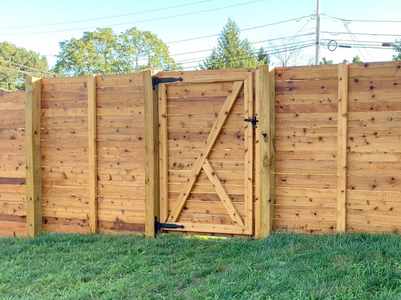 A wooden fence with vertical planks and a matching gate in the center, featuring black metal hinges and latch. Green grass is in the foreground, while freshly cleaned by SUDS Power Washing, trees and power lines are visible in the background.