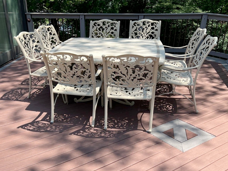 A white metal outdoor table with eight matching ornate chairs sits on a freshly cleaned wooden deck, thanks to SUDS Power Washing. Shadows from the intricate chair designs are cast on the deck, with green foliage visible beyond the railing.