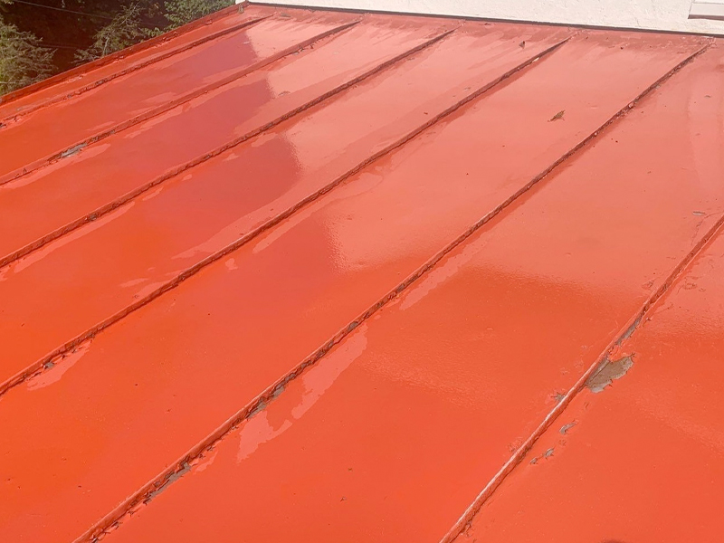 A close-up view of a red metal roof with vertical seams, showing some worn and chipped paint along the edges of the panels. The surface appears shiny and reflective in places, ideal for SUDS Power Washing to restore its original brilliance.