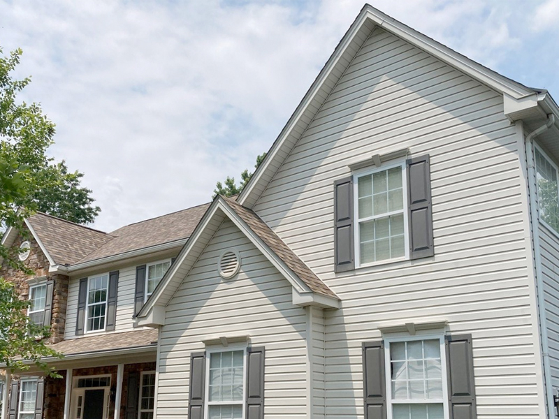 A two-story suburban house with light gray siding, white trim, and dark gray shutters under a partly cloudy sky. Trees are visible in the background, highlighting the spotless exterior after a SUDS Power Washing service.