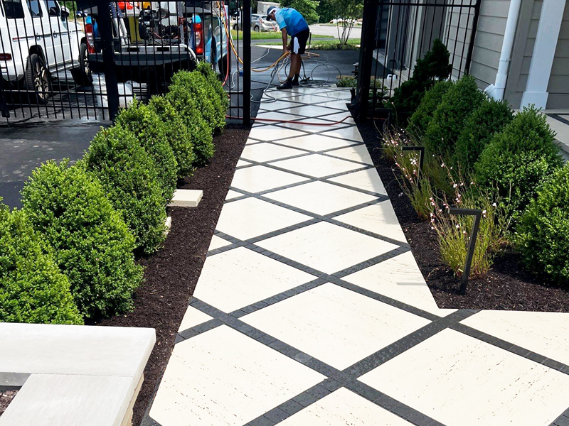 A person uses SUDS Power Washing to clean a patterned walkway with a hose. The cream surface with black crisscross lines is bordered by trimmed green bushes and dark mulch beds, with a black metal fence and parked vehicles in the background.
