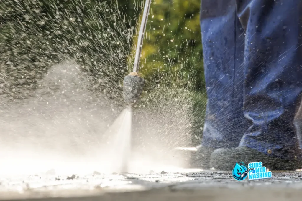 A person uses a pressure washer to clean an outdoor surface, with water spraying forcefully. The personโs legs and feet are visible, and the Suds Power Washing Services logo appears in the lower right corner.