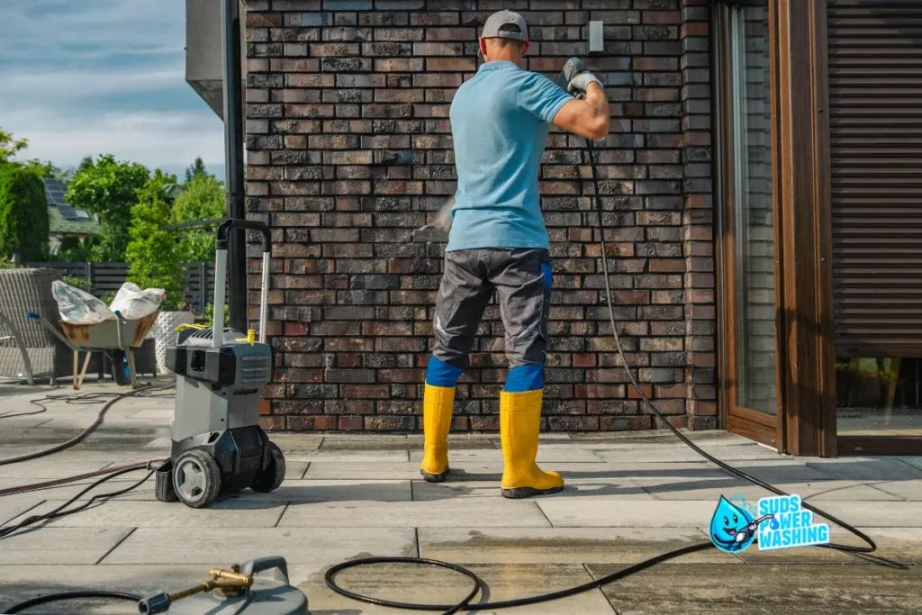 A person wearing yellow boots and a blue shirt is pressure washing a brick wall outside. Cleaning equipment and cords are on the patio, and the Suds Power Washer Near You logo appears in the lower right corner.