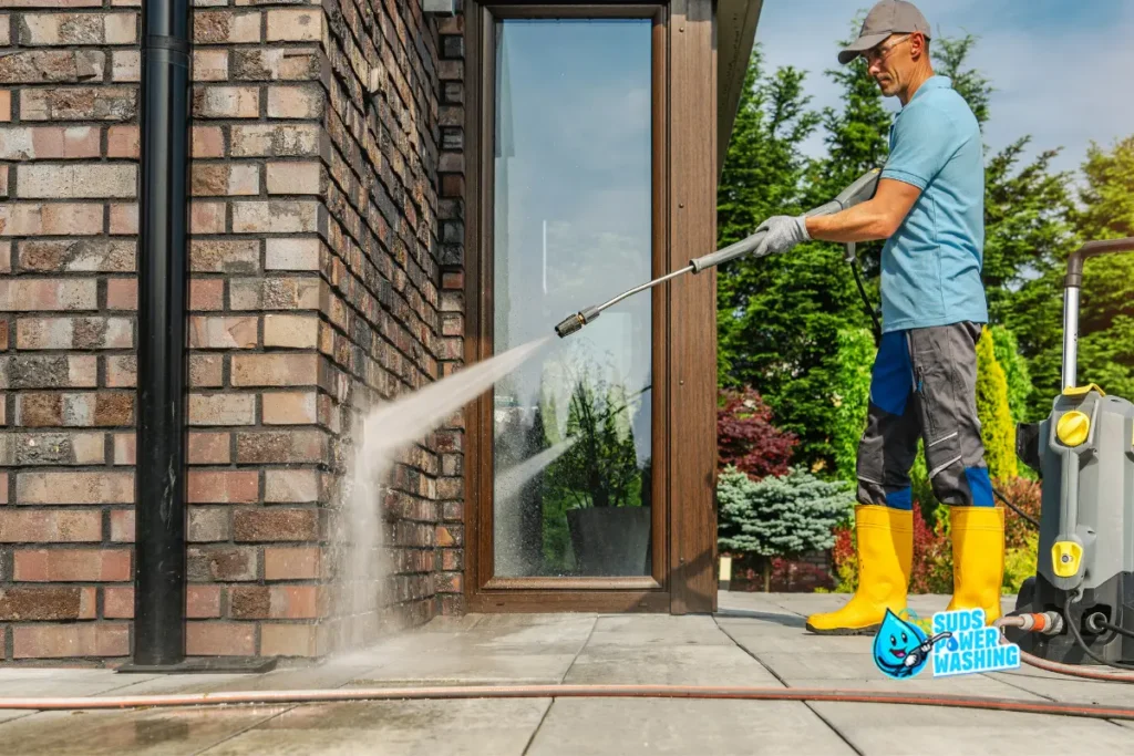 A man wearing yellow boots and gloves uses a power washer to clean a brick wall and patio outside a building, with trees and shrubs in the background. The Suds Power Washing logo appears in the lower right corner. what is power washing
