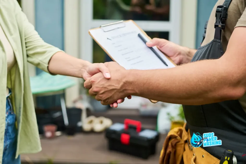 A person wearing work overalls shakes hands with another individual holding a clipboard, standing outside a house with tools and equipment visible in the background. Pressure Washing near me