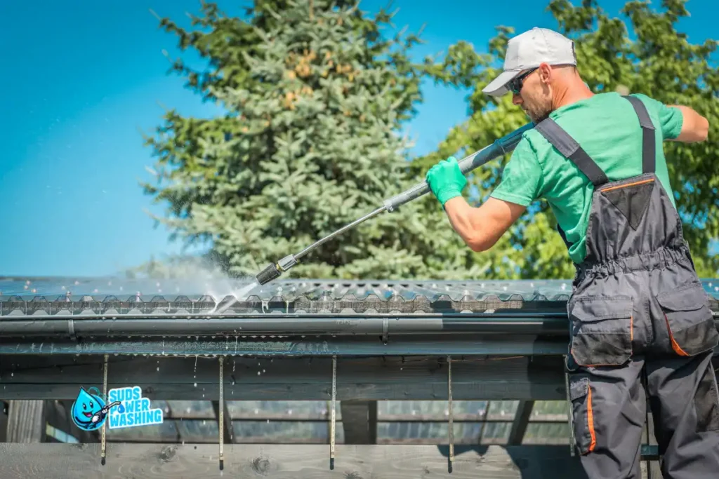 A man wearing gloves and overalls uses a pressure washer to clean the roof of a building, demonstrating expert soft washing techniques. Trees and a clear blue sky are in the background. There is a “Suds Power Washing” logo in the corner.