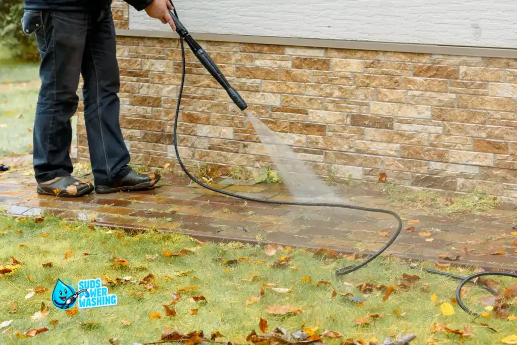 Person using a pressure washer to clean a brick walkway outside, with fallen leaves on the ground. A nearby brick wall and grass are visible, giving the scene a fresh, revitalized look. If you’re searching for “power washing near me,” this image perfectly showcases the results. The Suds Power Washing logo appears in the corner.