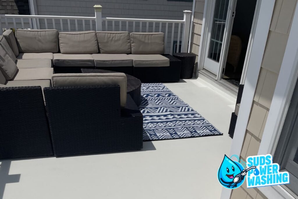 Outdoor patio with beige cushioned sectional sofa, patterned blue-and-white rug, and white railing. Suds Power Washing logo with a blue water droplet is in the lower right corner.