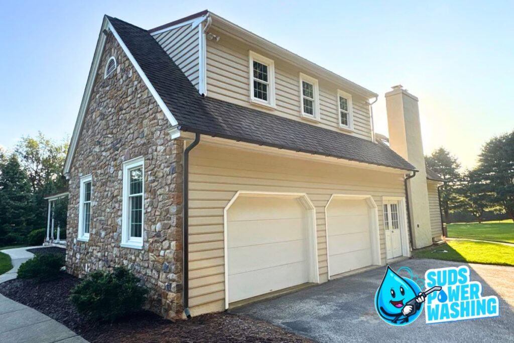 A two-car garage with light siding and stone accents is shown. The Suds Power Washing logo is in the corner. The driveway and yard are clean, with sunlight illuminating the house and green trees in the background.
