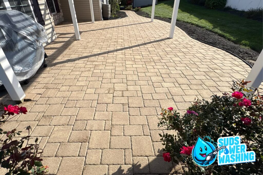 Clean, tan paver patio with white posts, rose bushes in the foreground, and sunlight casting shadows. “Suds Power Washing” logo with a blue water droplet character is in the lower right corner.
