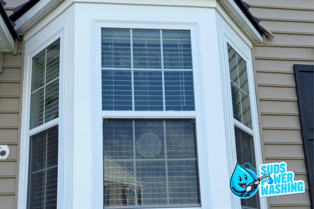 A clean bay window with white trim and closed blinds on a tan house. The Suds Power Washing logo with a blue water droplet is in the lower right corner, making this the go-to image for anyone searching for window cleaning near me or wanting to know how much does window cleaning cost.